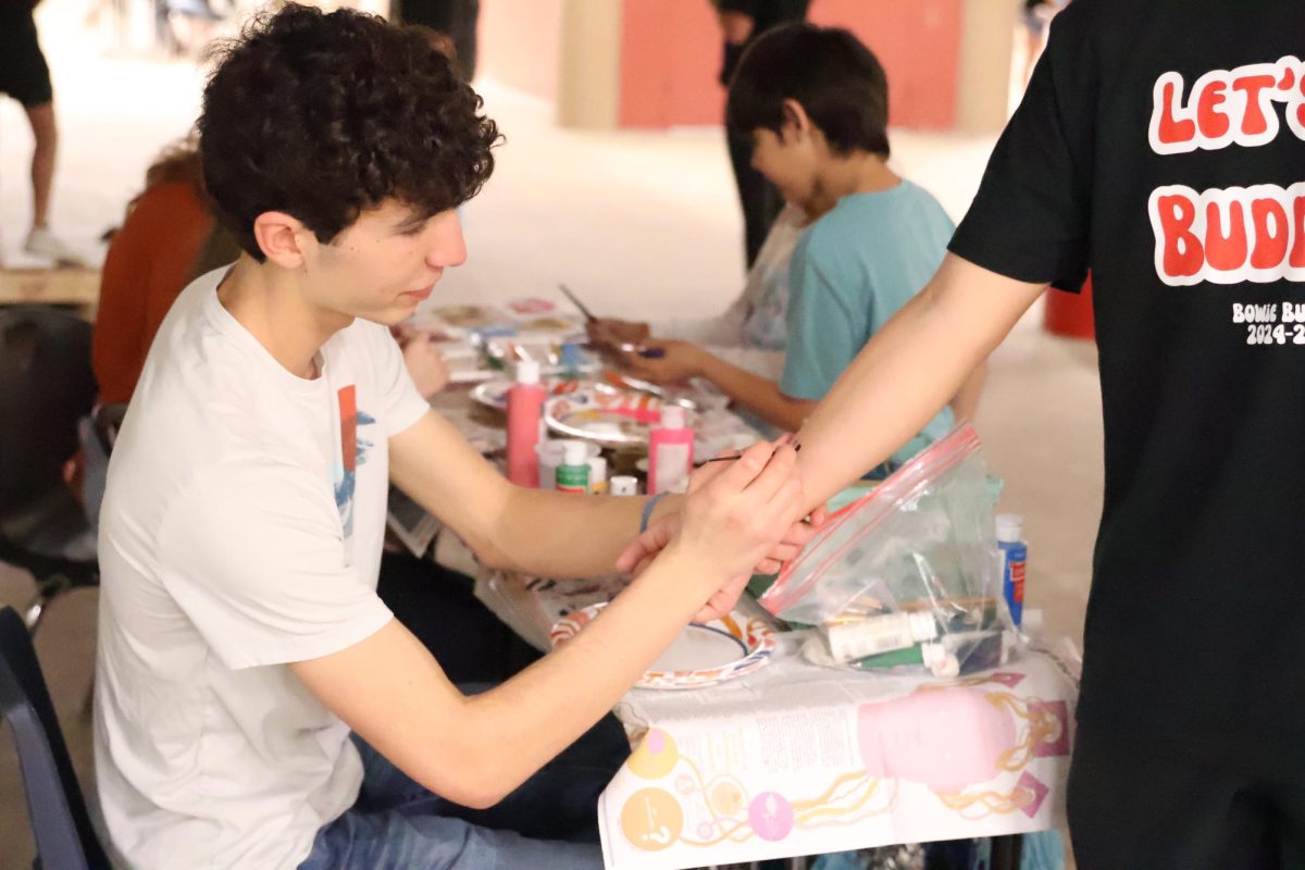 ARTISTIC BRILLIANCE: Painting with diligent strokes, junior Noah Dougherty creates a fun design on the arm of a fellow Bowie student. The Bowie Key Club held a painting booth at Bulldog Roundup offering candy and drawings to younger kids. “Bulldog Roundup was an incredibly fun and engaging experience,” Dougherty said. “I really enjoyed how it allowed us to connect with the community and showcase the various clubs at Bowie. It was a great way to interact with future Bowie students and share the spirit of our school.”
